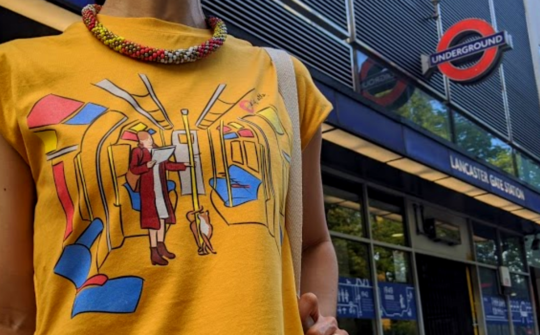 A tailored t-shirt dress in mustard yellow with Tubetiquette's 'Hold On' design outside a London Tube station. The design features a cat wrapping itself around a pole on the train.